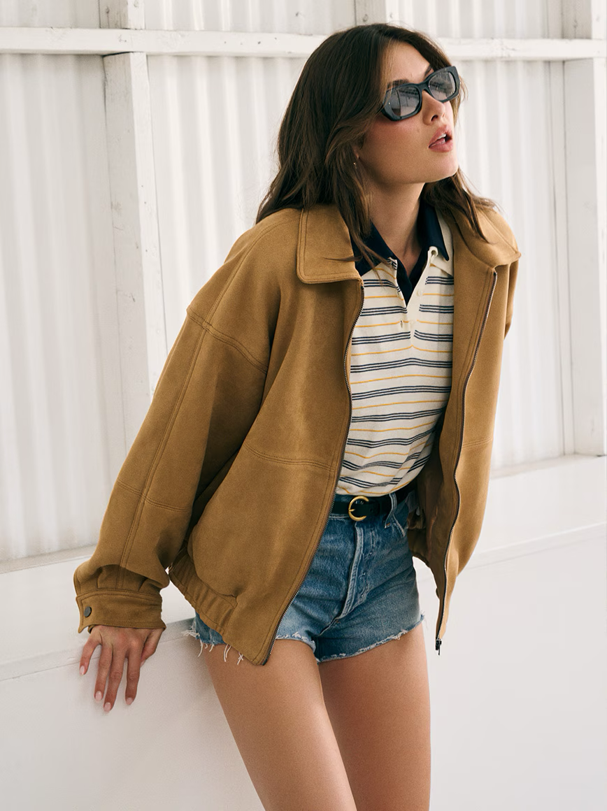 Person wearing sunglasses, brown jacket, striped shirt, and denim shorts.